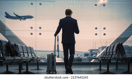 Airport Terminal: Businessman with Rolling Suitcase Walks, Uses Smartphone App for e-Business. Back View Silhouette of Traveling Man Waits for Flight in Boarding Lounge of Airline Hub with Airplanes - Powered by Shutterstock