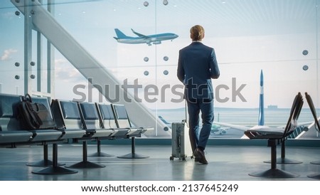 Airport Terminal: Businessman with Rolling Suitcase Walks, Uses Smartphone App for Internet e-Business. Traveling Entrepreneur Work Online while Waiting for a Flight in Boarding Lounge of Airline Hub
