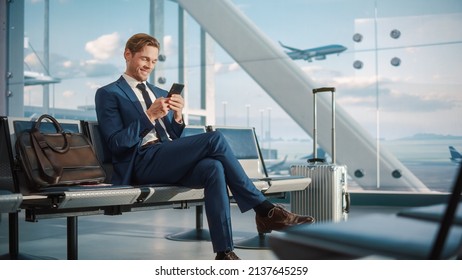 Airport Terminal Flight Wait: Smiling Businessman Uses Smartphone for e-Business, Browsing Internet with an App. Traveling Entrepreneur Work Online on Mobile Phone in Boarding Lounge of Airline Hub - Powered by Shutterstock