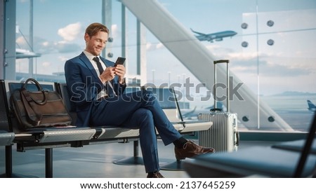 Airport Terminal Flight Wait: Smiling Businessman Uses Smartphone for e-Business, Browsing Internet with an App. Traveling Entrepreneur Work Online on Mobile Phone in Boarding Lounge of Airline Hub