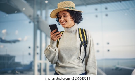 Airport Terminal: Happy Traveling Black Woman Looks Around Searching Flight Gates and Plane, Uses Smartphone, Checking Trip Destination on Internet. African American Female Wondering in Airline Hub
