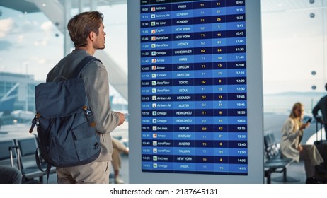 Airport Terminal: Young Man Looking At Arrival And Departure Information Display Looking For His Flight. Backgrond: Diverse Crowd Of People Wait For Their Flights In Boarding Lounge Of Airline Hub