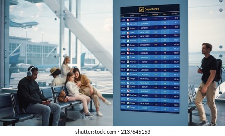 Airport Terminal: Young Man Looking at Arrival and Departure Information Display Looking for His Flight. Backgrond: Diverse Crowd of People Wait for their Flights in Boarding Lounge of Airline Hub