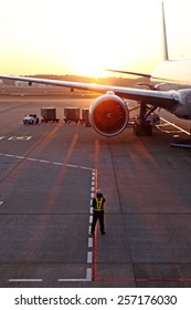 A Airport Traffic Controller Ground Crew Guiding An Airplane On An Airport Runway Against A Fiery Sunset.