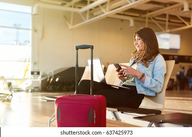 Airport Young female passenger on smart phone and laptop sitting in terminal hall while waiting for her flight
