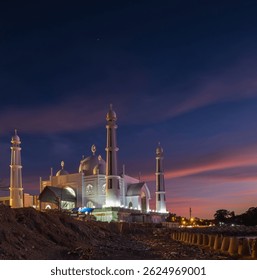 Mezquita Al Hakim en la playa de Padang, Sumatra Occidental, que brilla con minaretes blancos contra un cielo azul y rosa crepuscular, situado junto a la costa con farolas distantes, capturando la serena belleza costera.