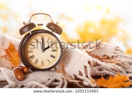 Alarm clock, acorns, scarf and autumn leaves on table outdoors. Daylight saving time end