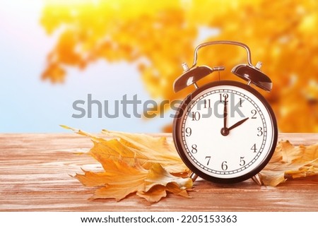Alarm clock and autumn leaves on table outdoors. Daylight saving time end
