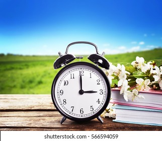 Alarm Clock With Books And Spring Blooming Branch On Wooden Table Against Landscape Background. Time Change Concept