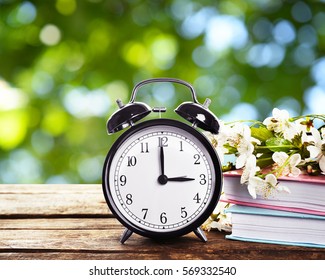 Alarm Clock With Books And Spring Blooming Branch On Wooden Table Against Foliage Background. Time Change Concept