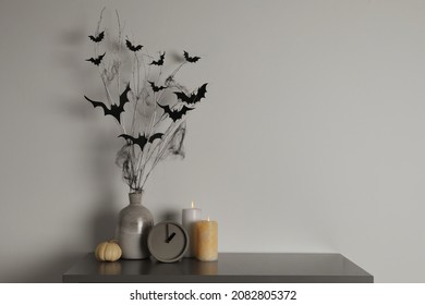 Alarm Clock, Burning Candles And Vase With Paper Bats  On Table Near White Wall, Space For Text. Halloween Decor