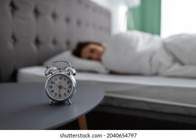 Alarm clock on bedside table and blurred view of millennial lady lying in bed, covered with warm blanket, unwilling to wake up, selective focus. Young woman feeling sleepy, free space