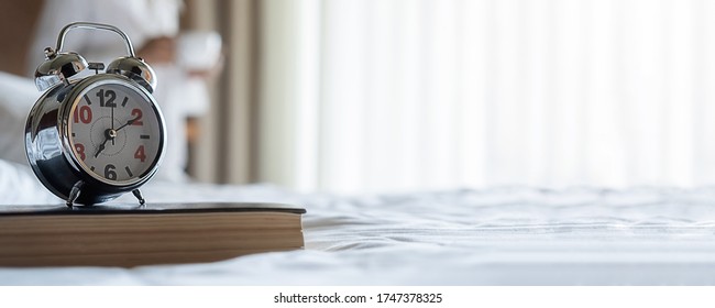 Alarm clock on book in white bedroom with woman drinking coffee after get up in morning sunrise background. Morning lifestyle concept.