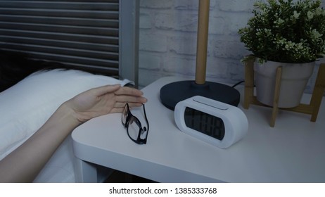 Alarm Clock On Nightstand Ringing In Midnight. Female Hand Taking Finding Eyeglasses On Table While Wake Up In Late Night In Dark Bedroom. Glasses And Plants In Home Interior On Desk By White Bed.