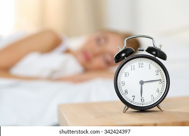 Alarm clock standing on bedside table going to ring early morning to wake up woman in bed sleeping in background. Early awakening, not getting enough sleep, oversleep, getting work time concept - Shutterstock ID 342424397