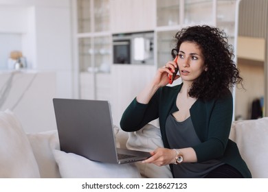 Alarmed Curly-haired Spanish Woman, Talking On The Phone, Sitting With A Laptop On The Couch With Her Eyes Open, Received Unexpected News, Upset. Remote Working Beautiful Spanish Businesswoman.