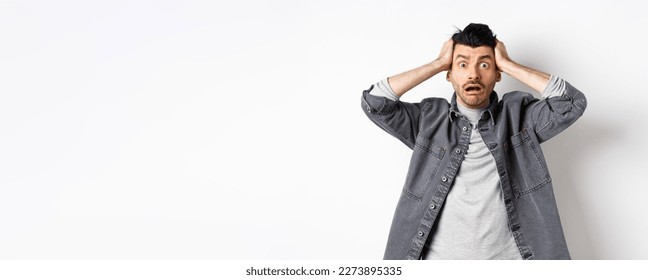 Alarmed young man grab head and pop eyes at camera, looking worried and troubled, having big problem, panicking on white background.