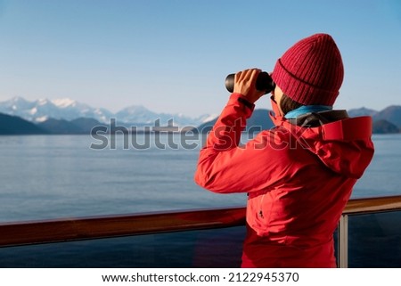 Alaska Glacier Bay cruise ship passenger looking at Alaskan mountains with binoculars exploring Glacier Bay National Park, USA. Woman on travel Inside Passage enjoying view. Vacation adventure