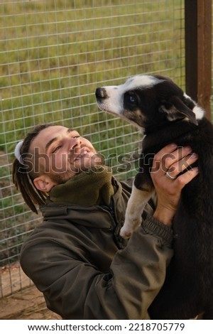 Alaskan Husky kennel, volunteer takes care of dogs. Young Caucasian man with dreadlocks chooses puppy from shelter, sitting in aviary, smiling and rejoicing. Human holds blue eyed puppy with hands.