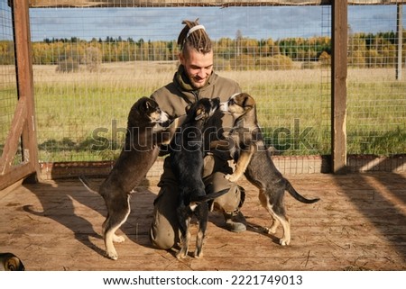 Alaskan Husky kennel, volunteer takes care of dogs. Young Caucasian man with dreadlocks chooses puppy from shelter, sitting in aviary, smiling and rejoicing. Concept of adoption of abandoned pets.