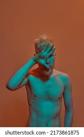 Albino Caucasian Guy Covering His Half Face And Looking At Camera. Young Modern Bearded Blonde Man Of Generation Z With Naked Torso. Isolated On Orange Background. Studio Shoot. Copy Space