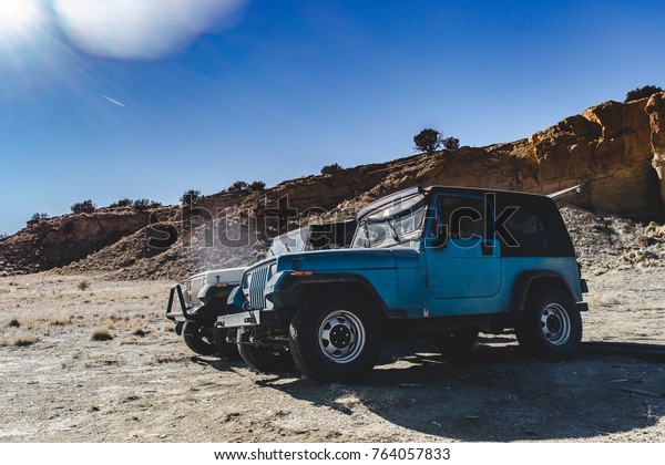 Albuquerque New Mexico 11 24 2017  Two jeeps\
side by side near a cliff in the desert with clear blue sky and jet\
stream in distance