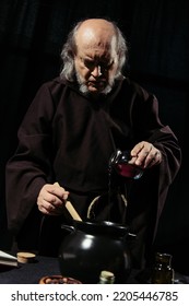 Alchemist In Dark Robe Pouring Liquid Into Pot While Cooking At Night Isolated On Black