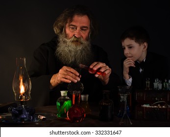 Alchemist Mixes Material From The Tube. Boy Watching Experience In The Laboratory. Magic, Mystery, A Fairy Tale. The Old Man In The Black. Sorcery