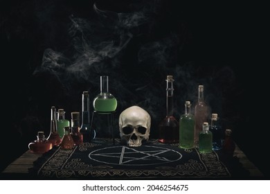 Alchemist , wizard or witch workplace. The altar. Magic potions and skull on the wooden table. Dark smoky background. Witchcraft and Halloween concept.