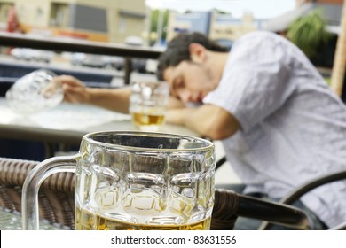 Alcohol Abuse: Drunk Young Man Or Student Lying Down On A Table With Beer Bock Still In Hand, Focus On Glass Up Front.