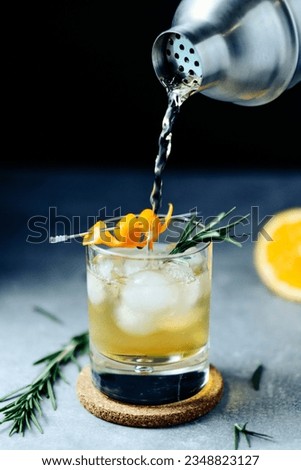 Alcohol cocktail pouring from shaker into glass with orange peel rosemary and ice on wooden desk