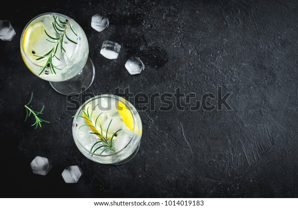 Alcohol drink (gin tonic cocktail) with lemon,\
rosemary and ice on rustic black stone table, copy space, top view.\
Iced drink with lemon and\
herbs.