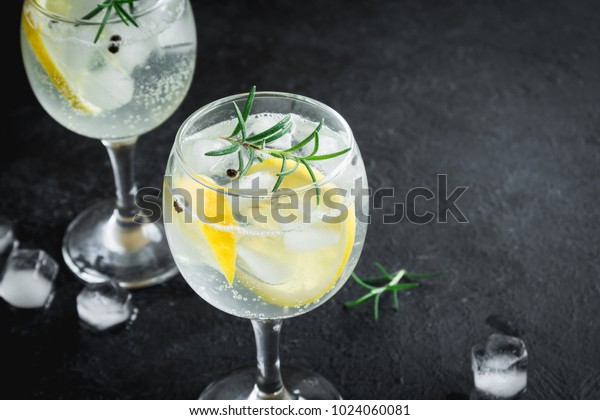 Alcohol drink (gin tonic cocktail) with lemon,\
rosemary and ice on rustic black stone table, copy space, top view.\
Iced drink with lemon.