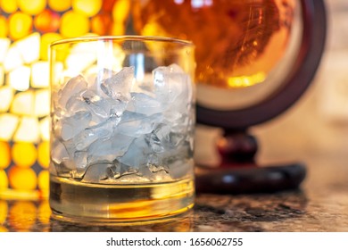 An Alcohol Glass With Ice Sits Aside A Round Container Of Scotch Whiskey On A Granite Countertop With Warm Light Glowing Behind.