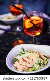 Alcoholic Cocktail With Ice And A Piece Of Orange And A Plate Of Caesar Salad With Chicken Breast And Parmesan On The Nightclub Table