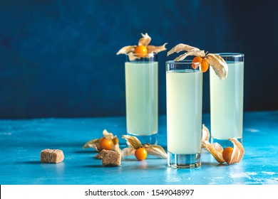 Alcoholic Cocktail With Ouzo Greek Anise Brandy Or Arabic Alcohol Drink Raki In Highball Glass, Decorated Physalis Peruviana And Brown Sugar On Blue Concrete Surface.