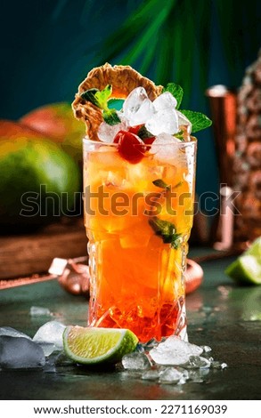Alcoholic cocktail with vodka, pineapple juice, mango, red syrup and ice. Long drink or summer cold mocktail. Tropical dark background with palm leaves and exotic fruits