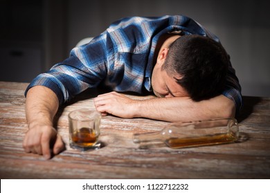 alcoholism, alcohol addiction and people concept - male alcoholic with glass of whiskey and bottle lying or sleeping on table at night