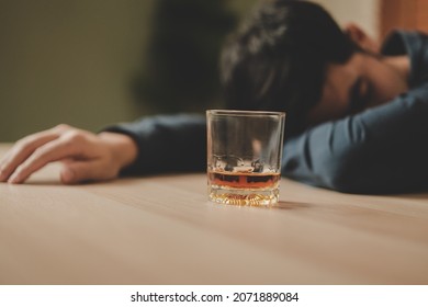 Alcoholism, Depressed Asian Young Man Sleep On Table While Drinking Alcoholic Beverage, Holding Glass Of Whiskey Alone At Night. Treatment Of Alcohol Addiction, Suffer Abuse Problem Alcoholism Concept