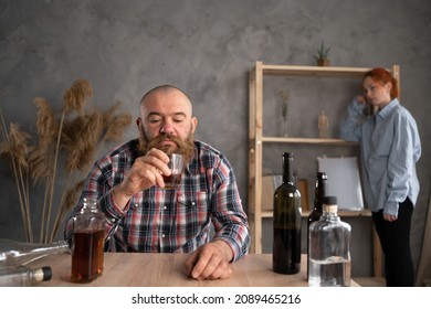 Alcoholism In The Family, A Sad Drunk Husband Pours Alcohol Into A Glass And A Sad Relaxed Wife In The Background, Alcohol Abuse At Home. Addiction And Depression. Copy Space.