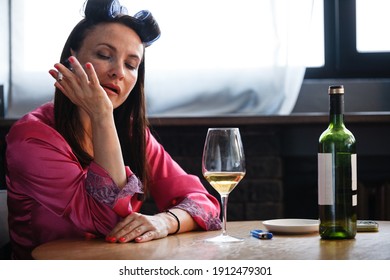 Alcoholism problem concept. Sad middle aged woman in stress sits in the kitchen, drinks white wine from a glass and smokes a cigarette. A depressed female is addicted to alcohol at home