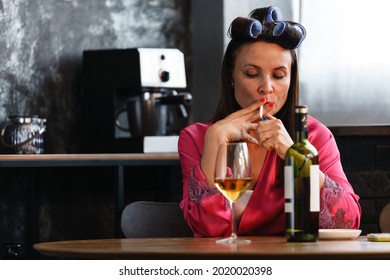 Alcoholism Problem Concept. Sad Middle Aged Woman In Stress Sits In The Kitchen, Drinks White Wine From A Glass And Smokes A Cigarette. A Depressed Female Is Addicted To Alcohol At Home