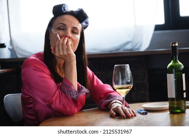 Alcoholism Problem Concept. Sad Middle Aged Woman In Stress Sits In The Kitchen, Drinks White Wine From A Glass And Smokes A Cigarette. A Depressed Female Is Addicted To Alcohol At Home