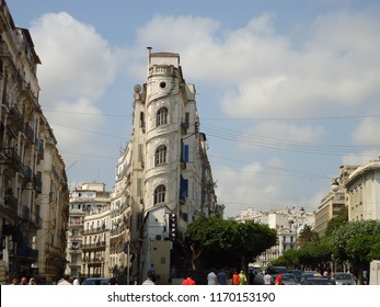 Algiers, Algeria. August 2018. Old French Building Of The Corner Shape Surrounded By Shorter Buildings And Trees And People Crossing The Road