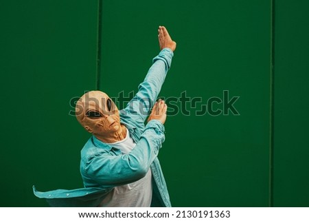 Alien doing dab posture and gesture against a green wall background. Extraterrestrial with human clothes. Concept of victory and satisfaction. Happiness and freedom immigration concept