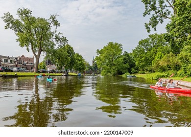 Alkmaar, North Holland, Netherlands. June 11, 2016. Scene In A Water Canal With Tourists Rowing Their Kayaks Against Cloudy Sky, Leafy Trees, Street With Parked Cars, Church Tower In The Background