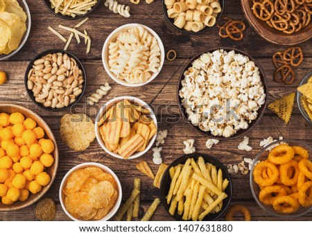 All classic potato snacks with peanuts, popcorn and onion rings and salted pretzels in bowl plates on wood. Twirls with sticks and potato chips and crisps with nachos and cheese balls. Top view