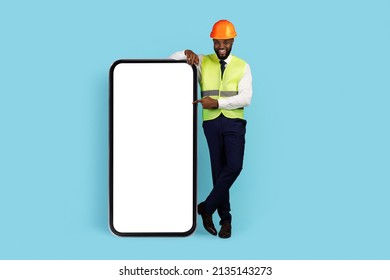 All For Construction. Happy Black Engineer Pointing At Big Blank Smartphone Screen, African American Man In Safety Vest And Hard Hat Advertising Mobile App, Standing Over Blue Background, Mockup