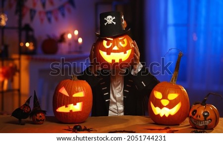 All hallows eve. Man in  skeleton costume and hat holding glowing jack o lantern carved pumpkin in front of his face, standing behind table with Halloween decorations in dark room