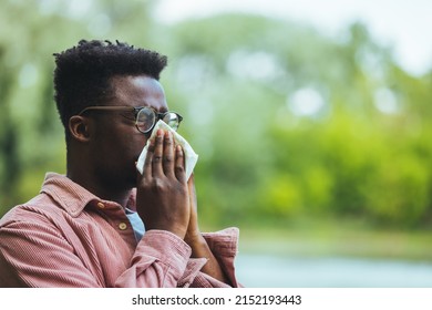 Allergic black man blowing on wipe in a park on spring season. Man with allergy or cold, blowing his nose with a tissue, looking miserable unwell very sick, isolated outside green trees background.  - Powered by Shutterstock